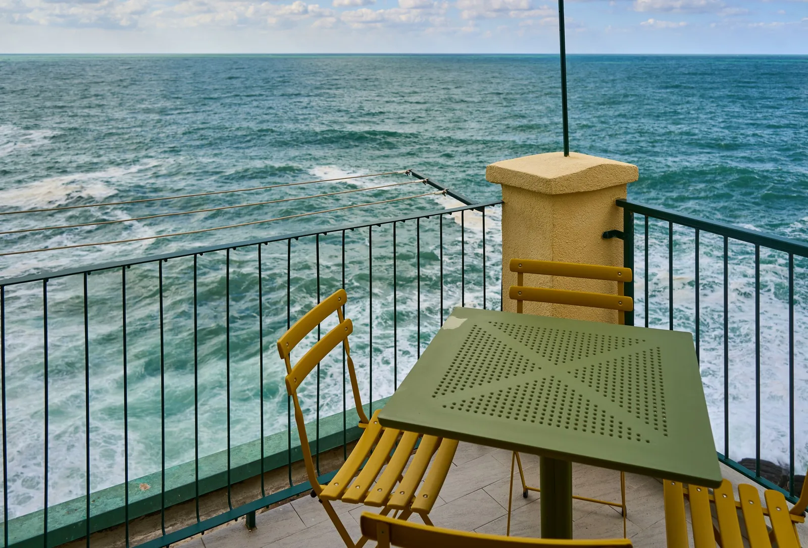 A yellow bistro table and chairs on a balcony with a metal railing overlooking the sea