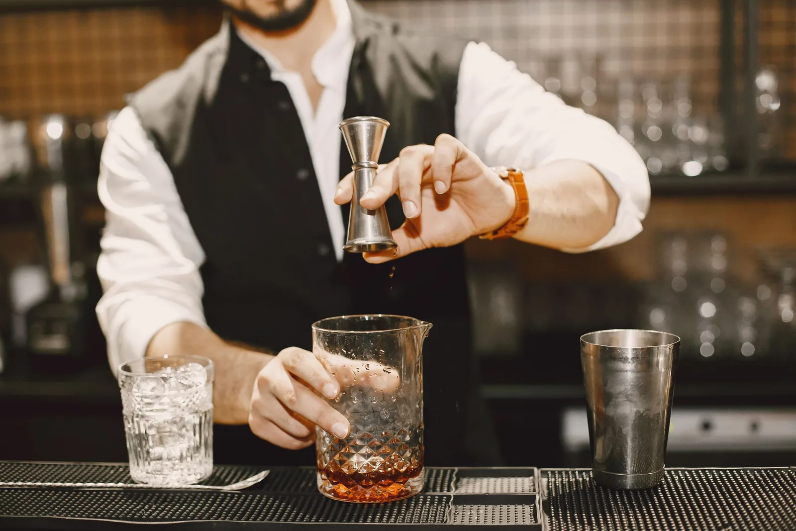 A bartender in a waistcoat measuring spirits with a jigger over a mixing glass at a dark bar