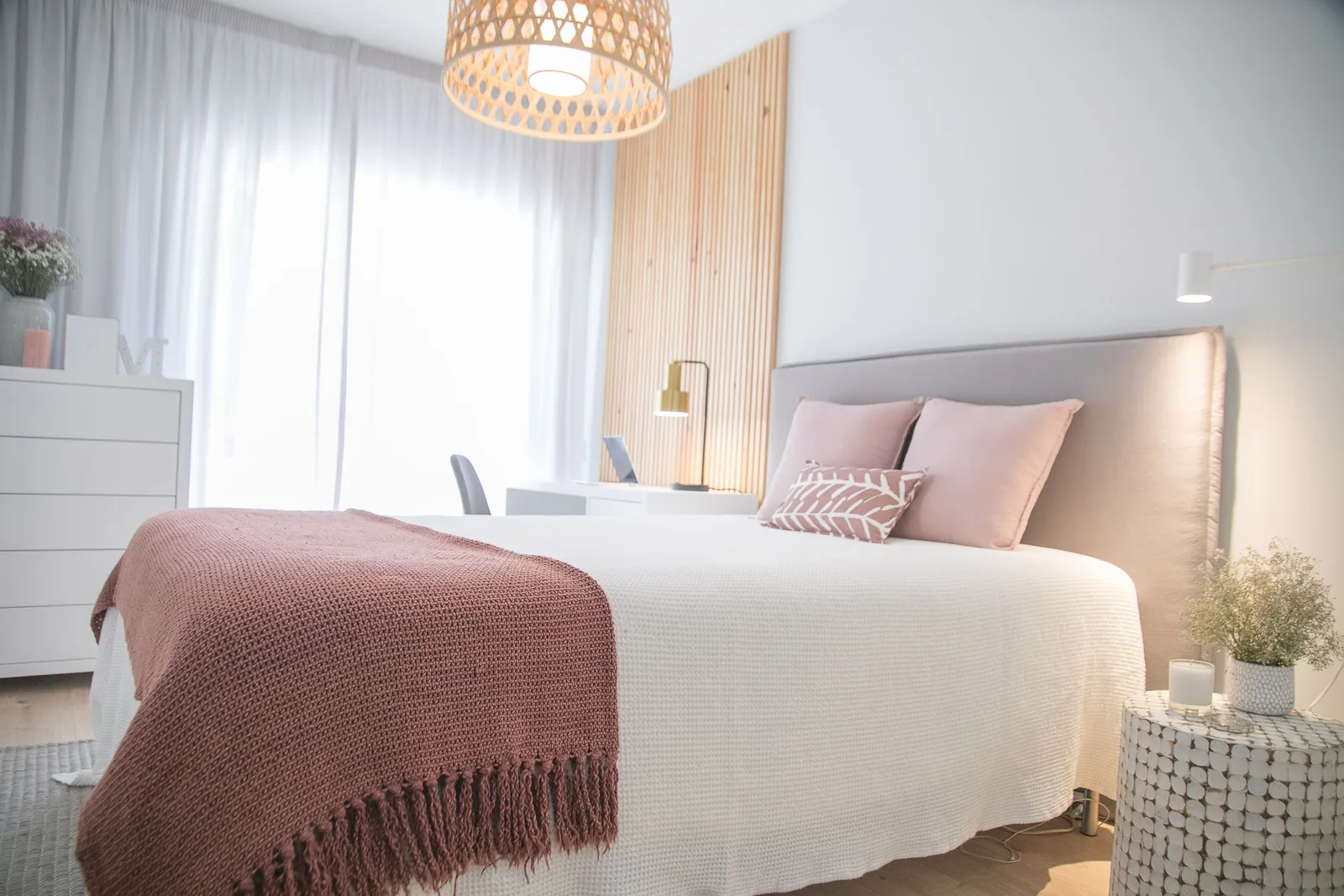 A bright bedroom with a white bed, a pink throw, sheer curtains and a woven pendant lamp