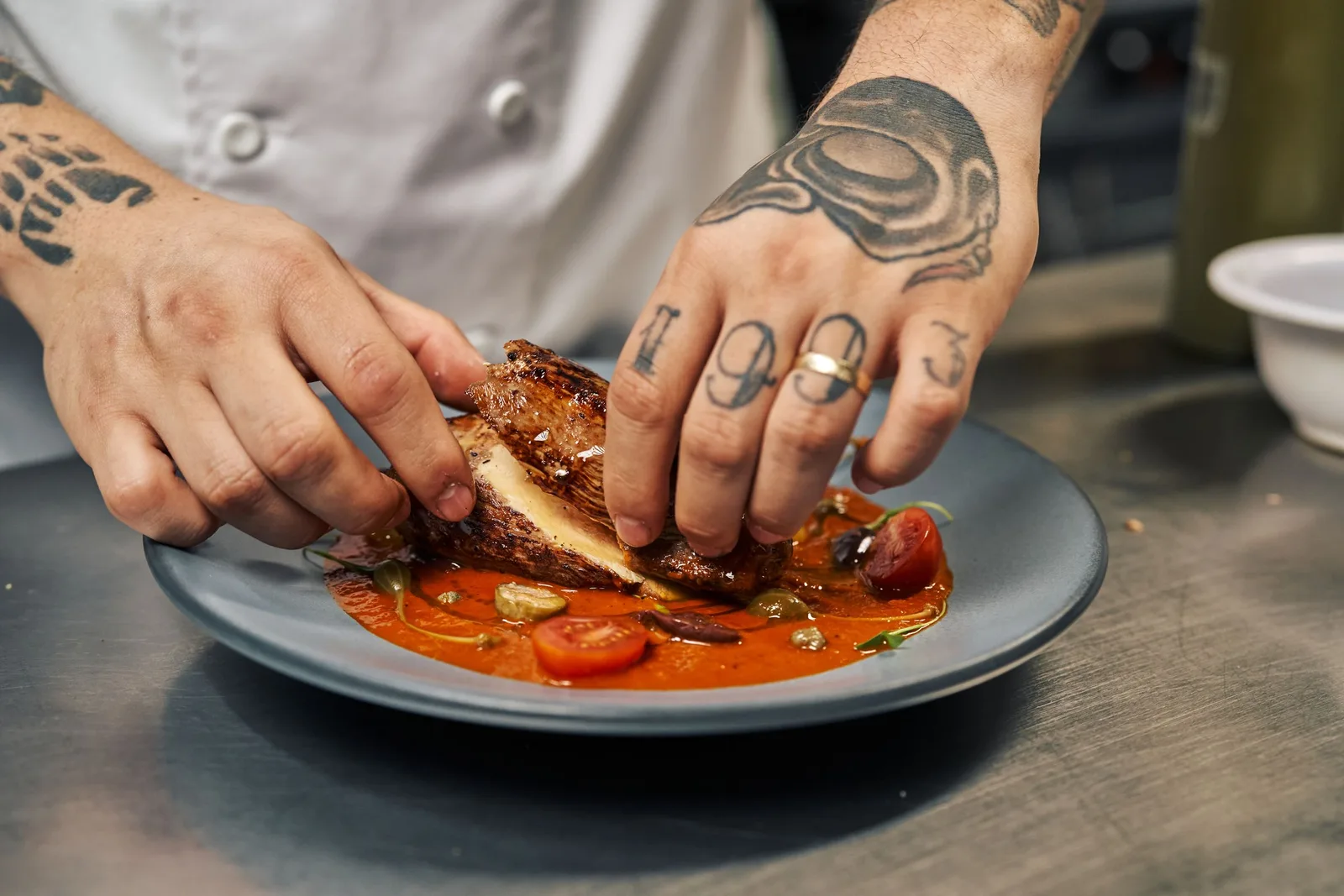 A chef’s hands placing roasted chicken and tomatoes onto a dark plate