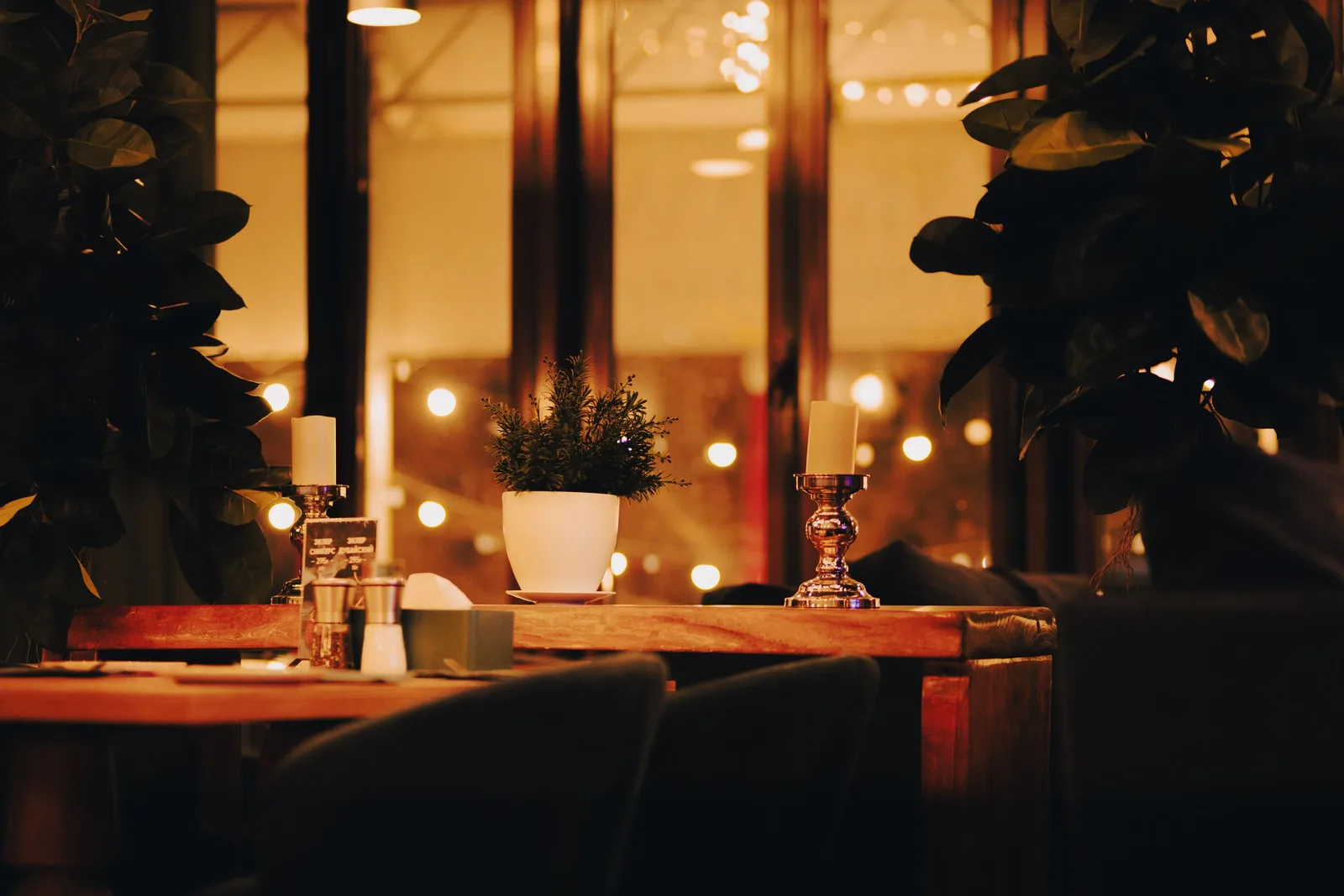 A restaurant table by a window at night, set with candles, a small potted plant and warm string lights behind