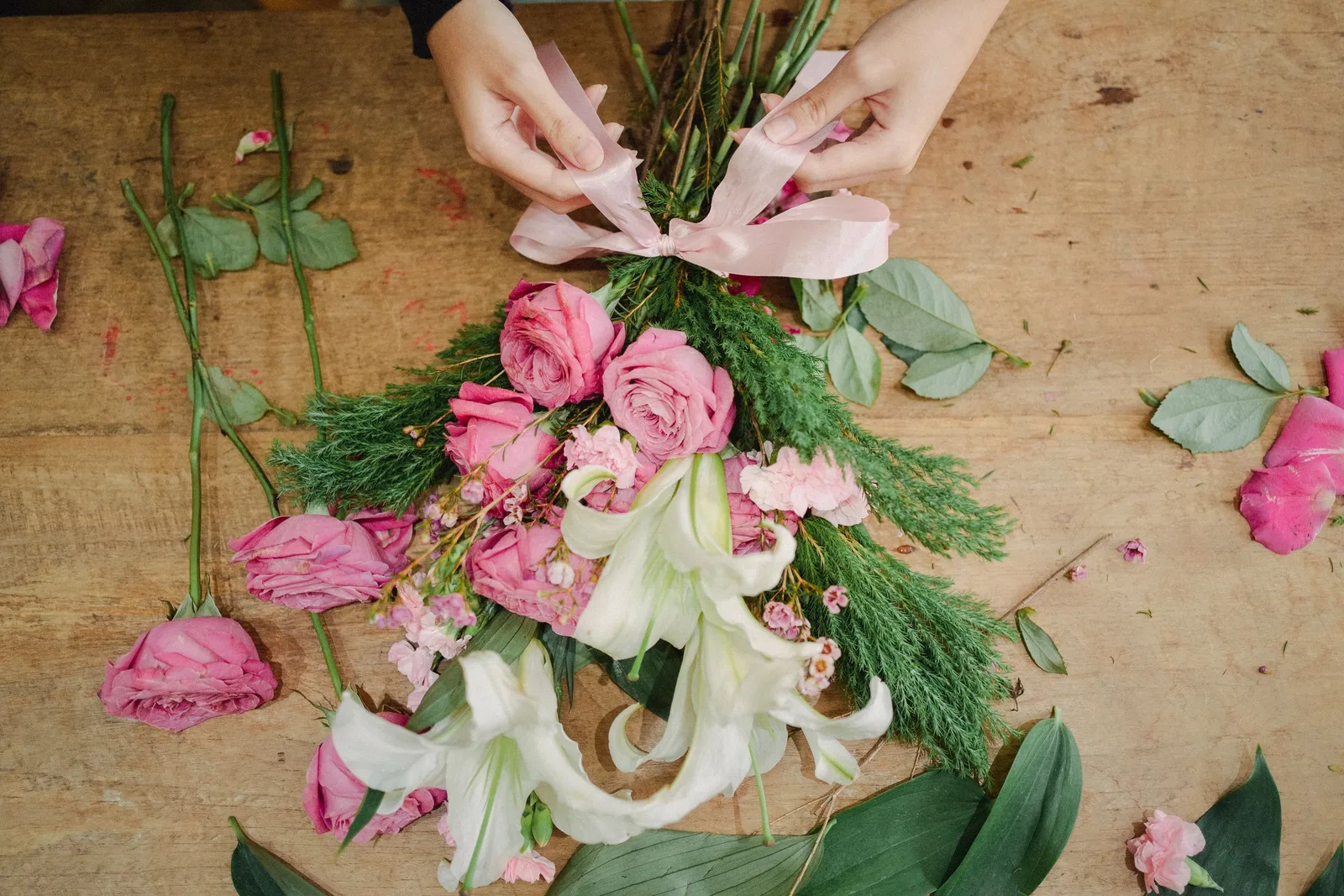 Overhead view of hands tying a ribbon around a bouquet of pink roses and lilies on a wooden table