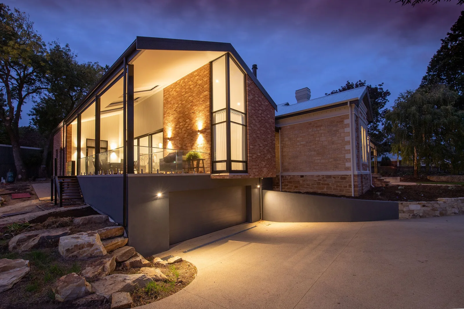 A modern glass and brick house at dusk with its interior lights glowing and a lit concrete path in front