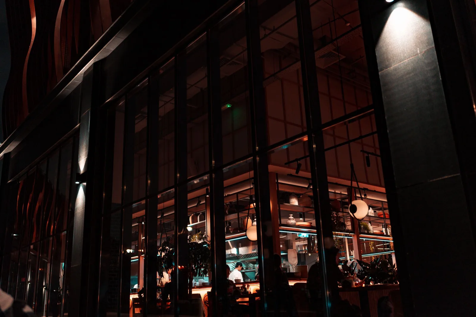 Tall lit windows of a restaurant seen from the dark street at night, with diners visible inside