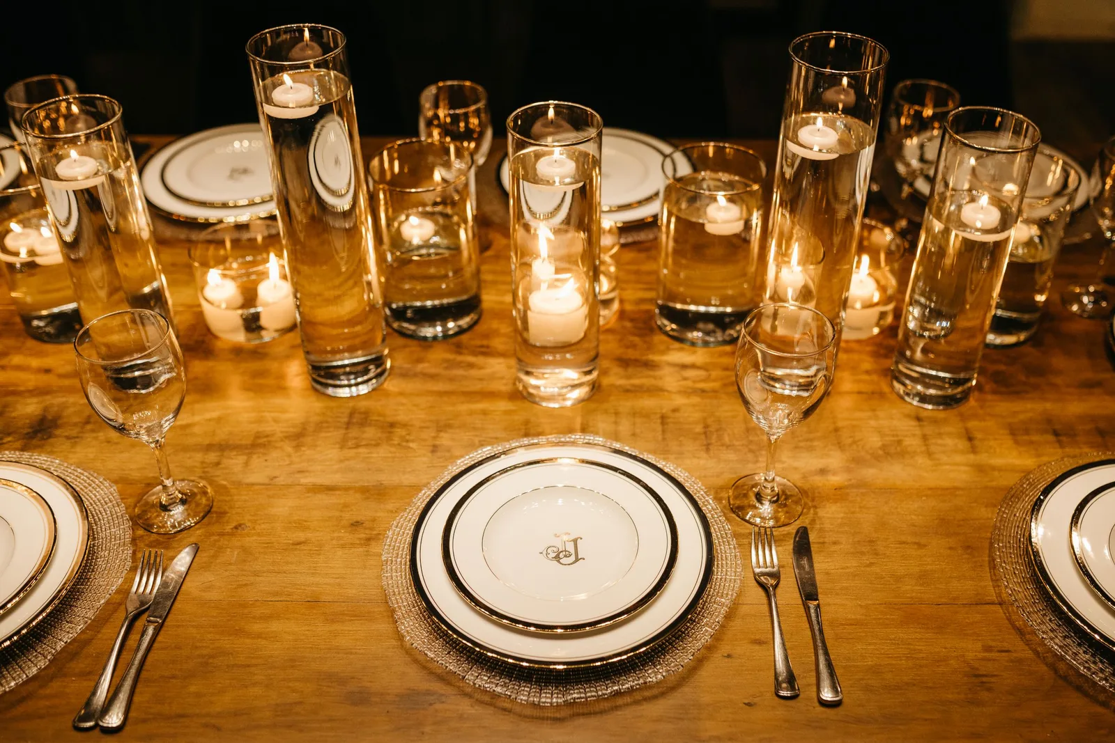 A long wooden dinner table set with white plates, glassware, cutlery and rows of lit candles in glass holders