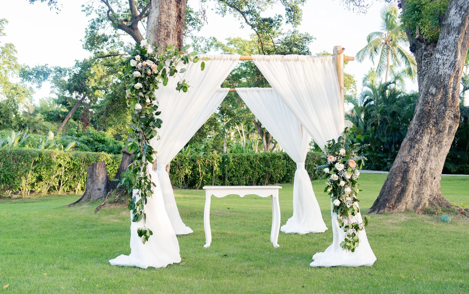 A white-draped wedding arch decorated with flowers and greenery on a lawn under large trees