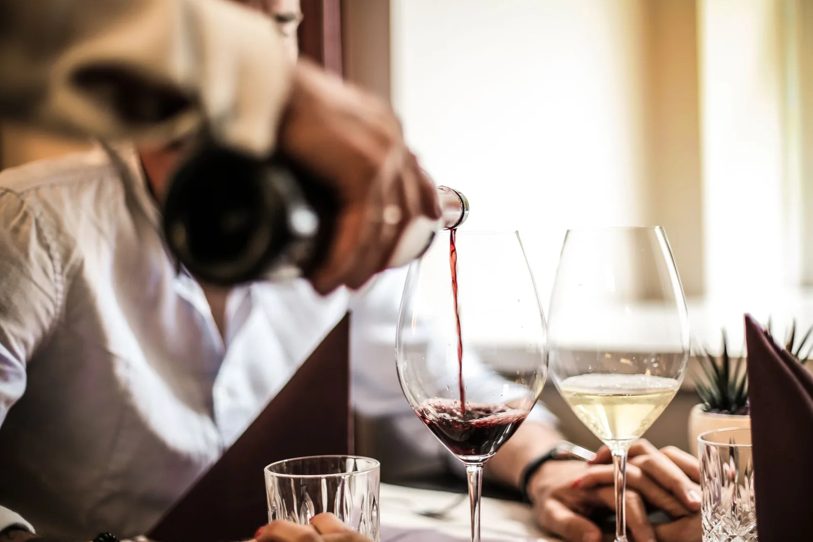 A waiter in a white shirt pouring red wine into a glass at a restaurant table
